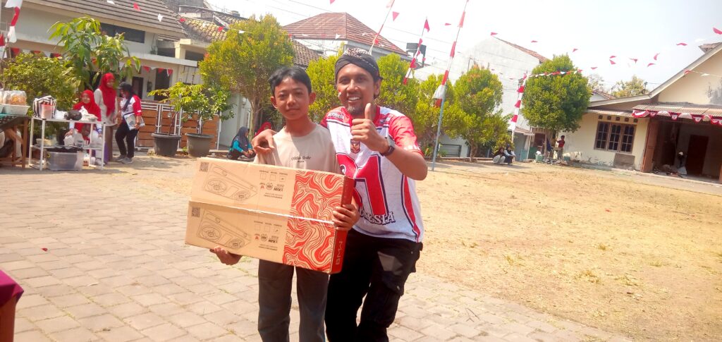 IMG_20260816_101903 - Mabur.co Two men pose for a photo at an outdoor festival, one wearing a red-and-white shirt giving a thumbs up while holding a large boxed item.