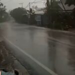 Residential street flooded with standing water during heavy rain; power lines and trees in the background.