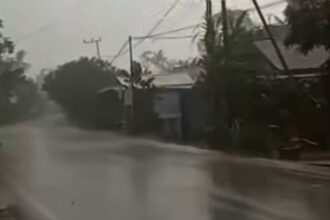 Residential street flooded with standing water during heavy rain; power lines and trees in the background.