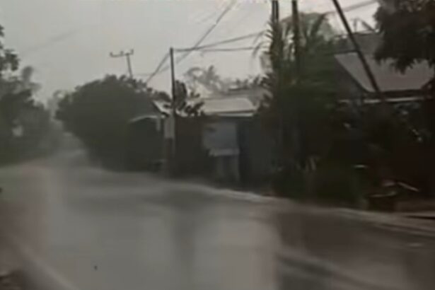 Residential street flooded with standing water during heavy rain; power lines and trees in the background.