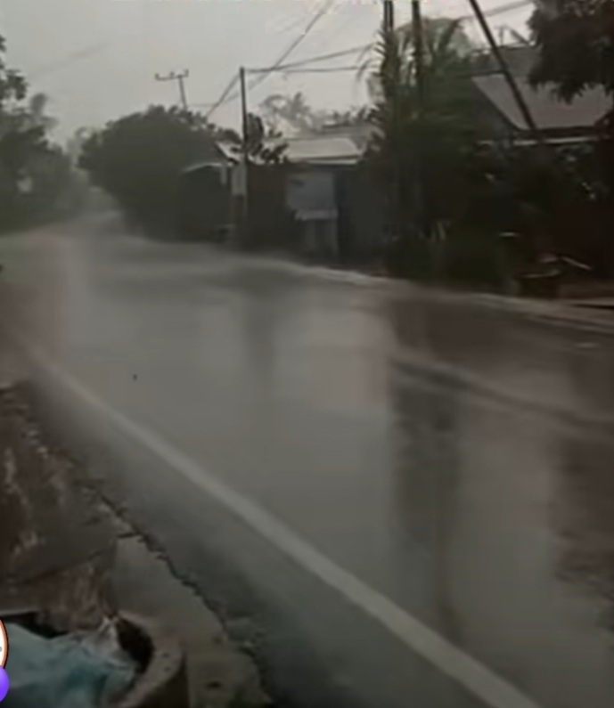 Residential street flooded with standing water during heavy rain; power lines and trees in the background.