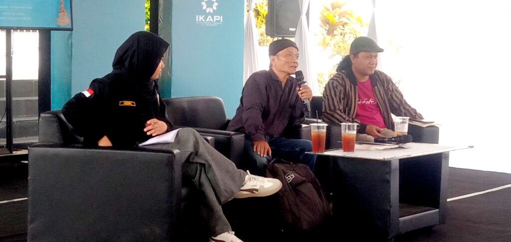 Panel of three people seated on black sofas, one person speaking into a microphone while others listen, with drinks on a table in front.