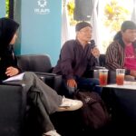 Panel of three people seated on black sofas, one person speaking into a microphone while others listen, with drinks on a table in front.