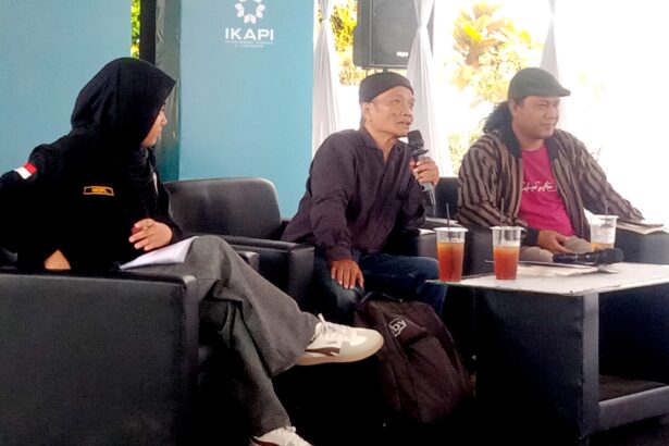 Panel of three people seated on black sofas, one person speaking into a microphone while others listen, with drinks on a table in front.