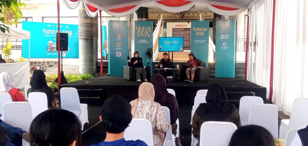Panel discussion on a stage with three speakers under red-and-white drapery and blue banners reading'Pameran Arsip' behind them
