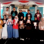 Group of students and organizers on a stage at a book fair, each holding a booklet or program.