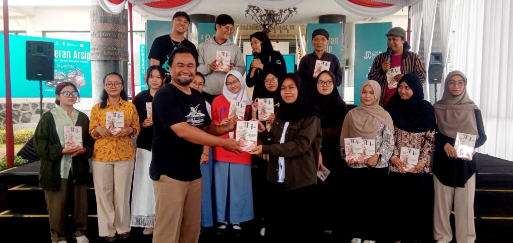 Group of students and organizers on a stage, smiling, as a man shakes hands with a student and hands her a booklet.