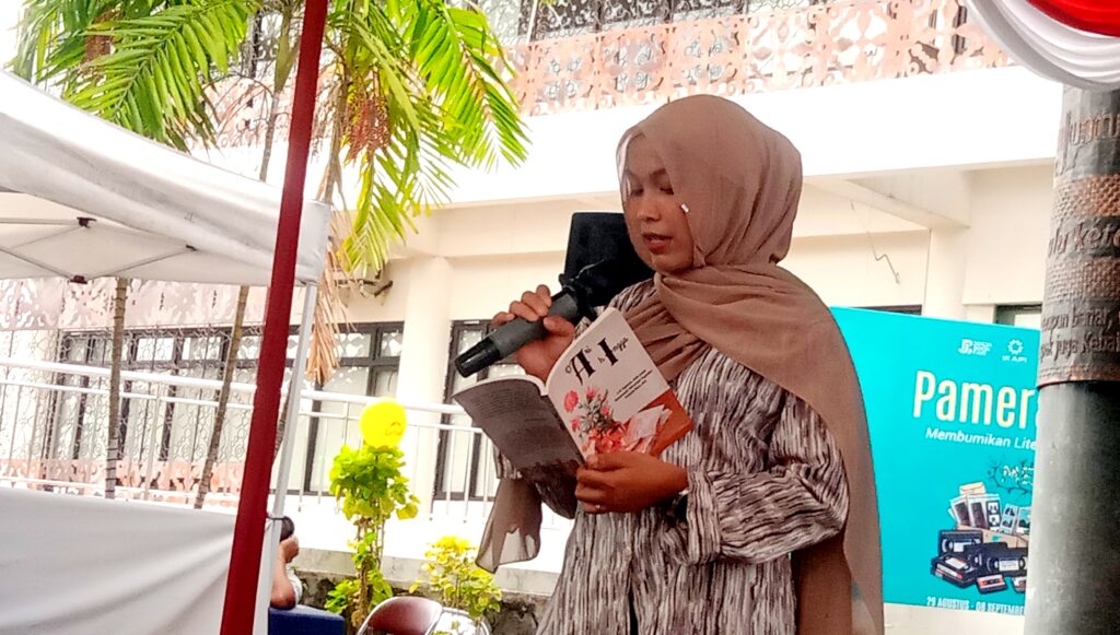 Woman wearing a brown hijab speaks into a microphone while reading from a pamphlet at an outdoor event, with a blue banner in the background.