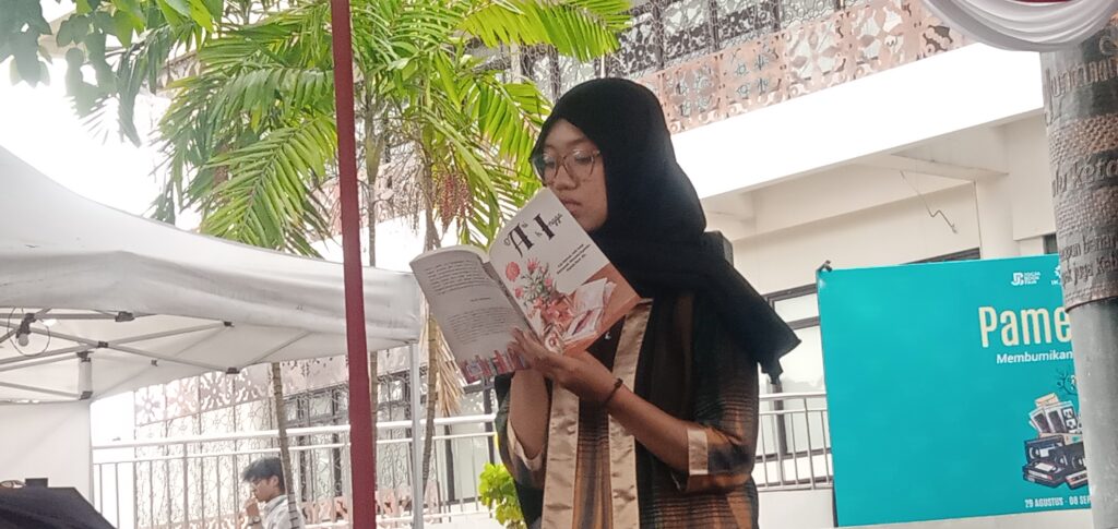 Woman wearing a black hijab reads a magazine outdoors beside palm trees and a blue poster board.