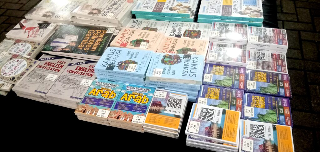 Stacks of language dictionaries and phrasebooks on a market table, including Indonesian-English and Arabic/Korean guides.