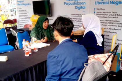 Woman in a green hijab chats with a man in a blue suit and a woman in a white hijab at a literacy booth with posters behind and water bottles on the table.
