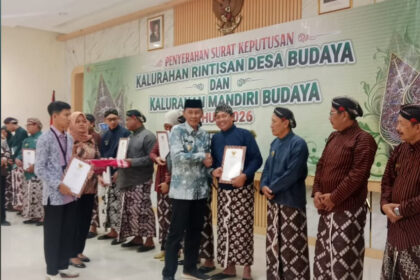 Recipients receive certificates at a cultural village awards ceremony, with a banner in Indonesian in the background.