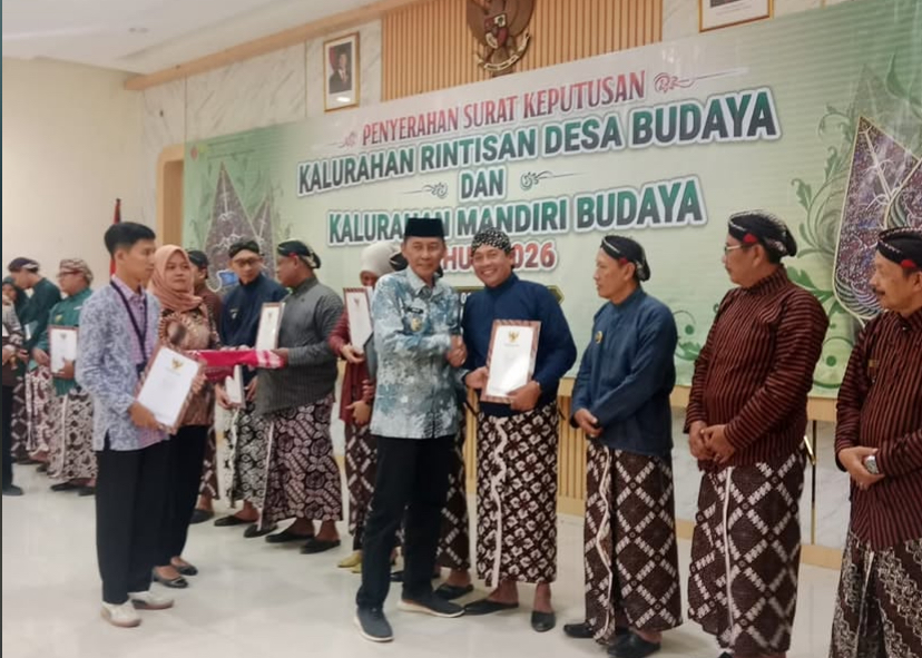 Recipients receive certificates at a cultural village awards ceremony, with a banner in Indonesian in the background.