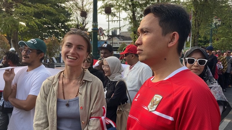 Smiling woman with sunglasses on her head stands beside a man in a red jersey at a crowded street event.