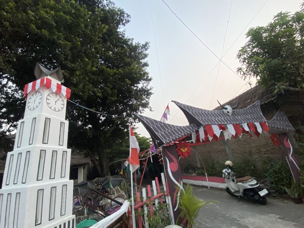 White decorative clock tower prop with red-and-white striped roof beside a festival street with bunting and a scooter nearby
