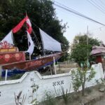 Decorated parade boat or float with red and white fabric on a residential street, trees in the background and a white wall fence nearby