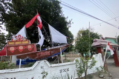 Decorated parade boat or float with red and white fabric on a residential street, trees in the background and a white wall fence nearby