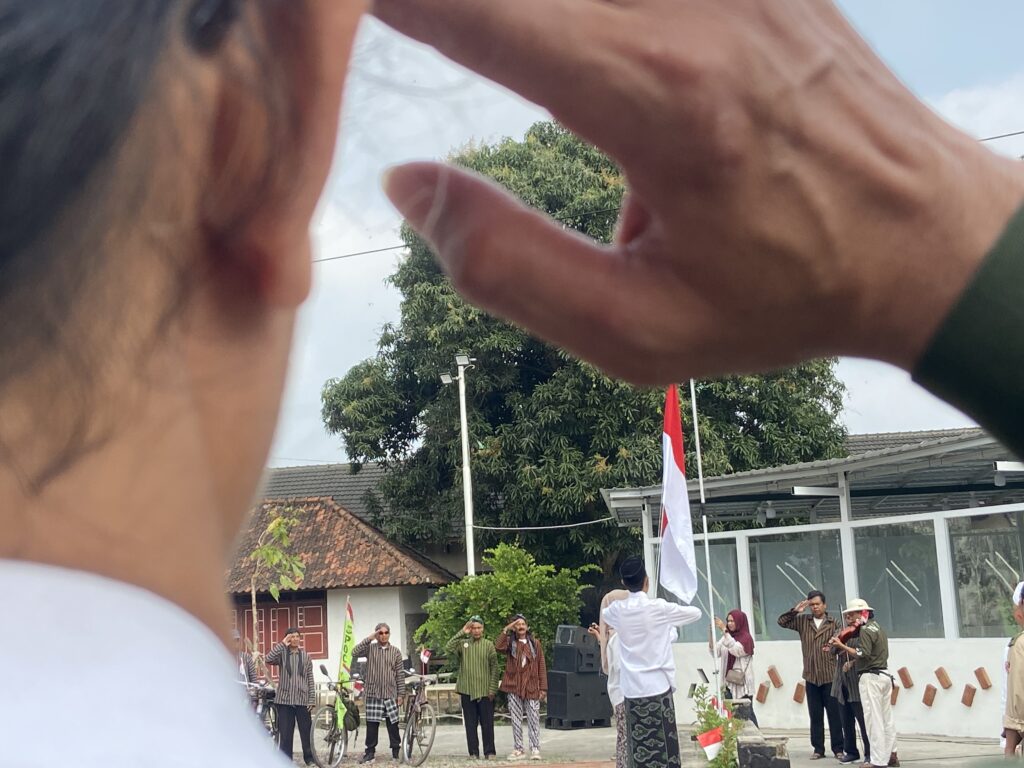 People in brown striped uniforms standing in a line outdoors, saluting during a flag-raising ceremony with Indonesian flags visible in the background.