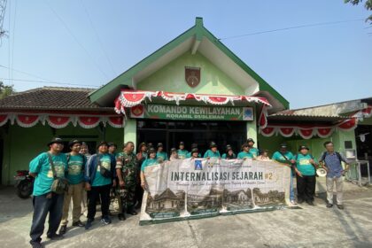 Group of people in teal shirts posing with a large banner in front of a green building with a peaked roof and red-white bunting decorations.",