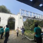 Group of people in teal shirts standing outdoors beside a white arched wall and a metal truss structure overhead on a sunny day; one person is speaking with a megaphone nearby.