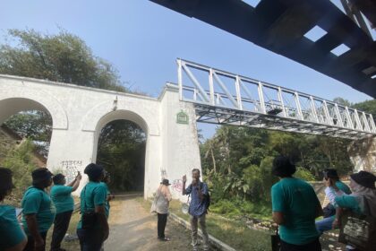 Group of people in teal shirts standing outdoors beside a white arched wall and a metal truss structure overhead on a sunny day; one person is speaking with a megaphone nearby.