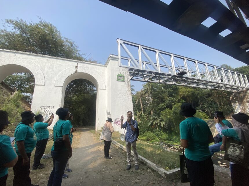 Group of people in teal shirts standing outdoors beside a white arched wall and a metal truss structure overhead on a sunny day; one person is speaking with a megaphone nearby.