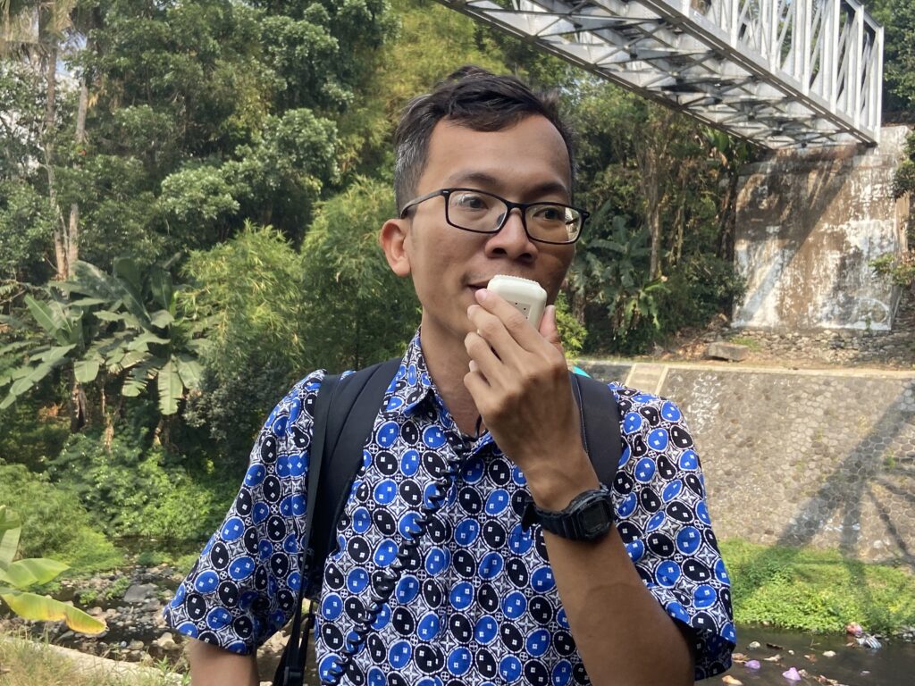 Man wearing glasses and a blue patterned shirt speaks into a handheld device outdoors, with greenery and a metal bridge in the background.
