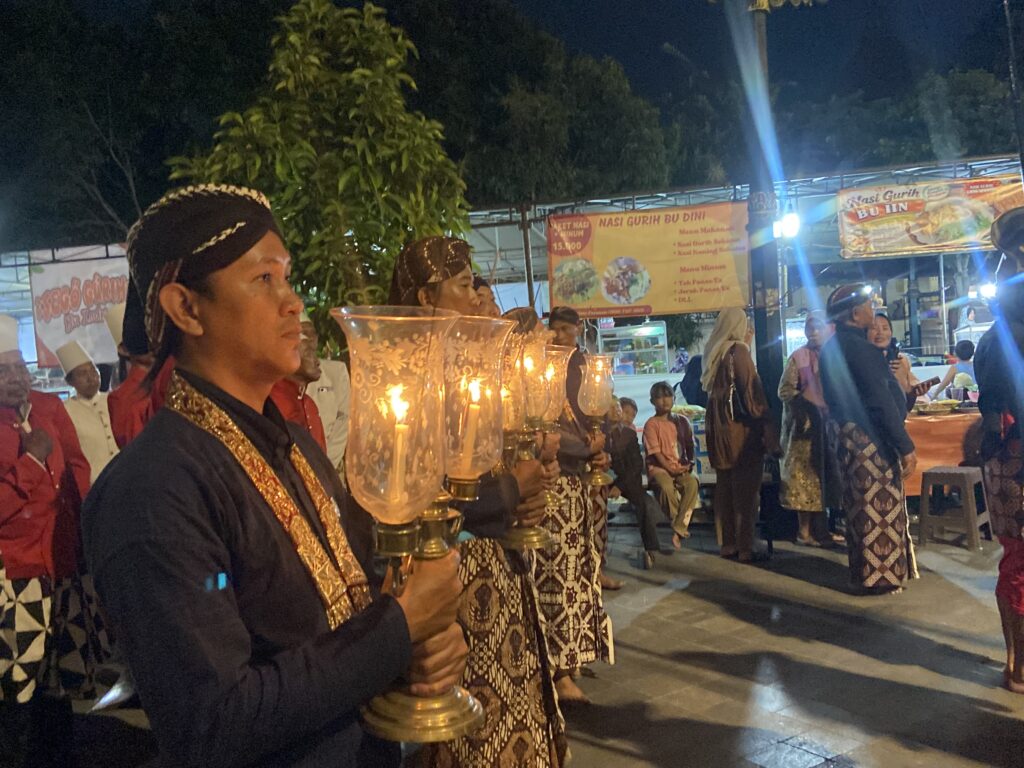 Group of people in batik outfits carrying illuminated lanterns during a nighttime ceremonial procession at a street market.