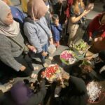 Group of people gathered around baskets of red tomatoes and flowering arrangements on a night street market.