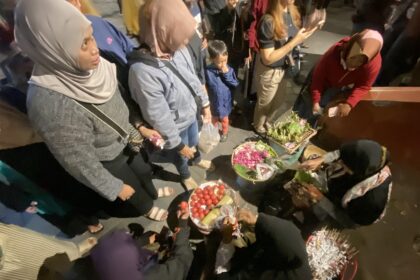 Group of people gathered around baskets of red tomatoes and flowering arrangements on a night street market.