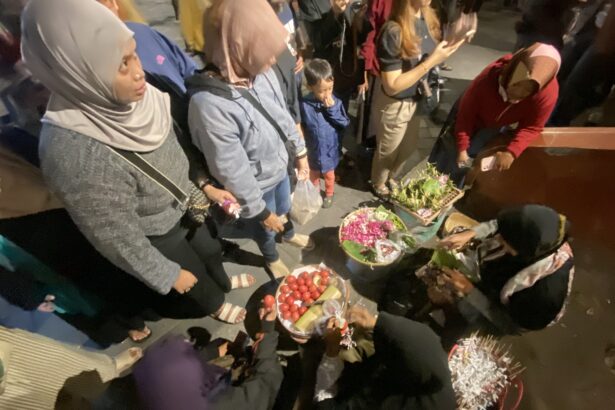 Group of people gathered around baskets of red tomatoes and flowering arrangements on a night street market.