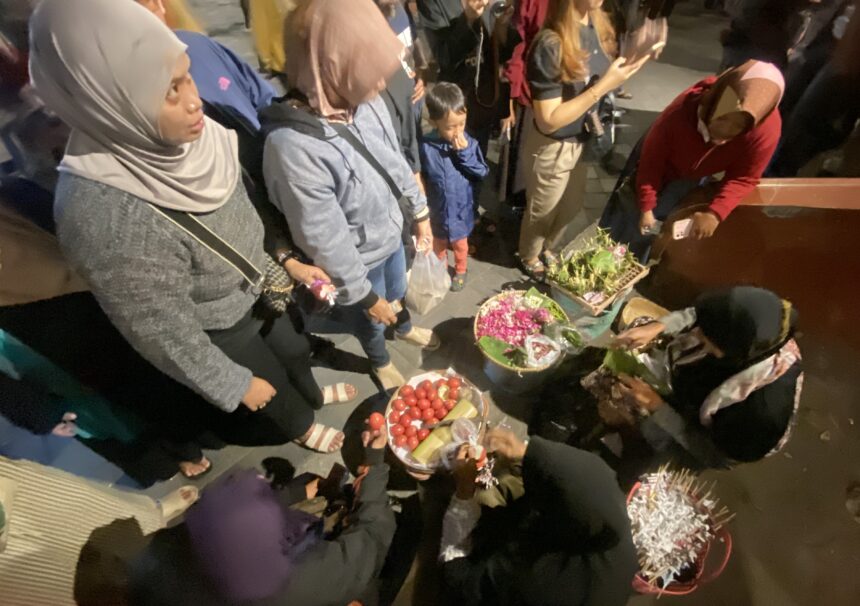 Group of people gathered around baskets of red tomatoes and flowering arrangements on a night street market.