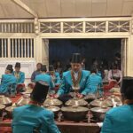 Group of musicians in turquoise traditional outfits playing percussion instruments in an indoor hall.