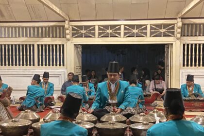 Group of musicians in turquoise traditional outfits playing percussion instruments in an indoor hall.