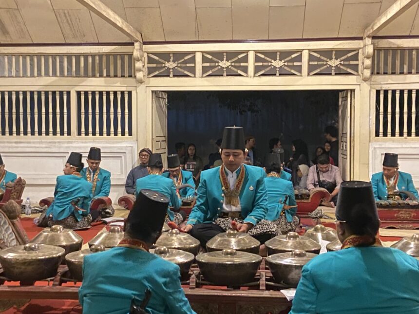 Group of musicians in turquoise traditional outfits playing percussion instruments in an indoor hall.