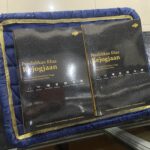 Two black hardcover books with gold Indonesian titles resting on a blue padded mat with a gold trim and patterned covers.