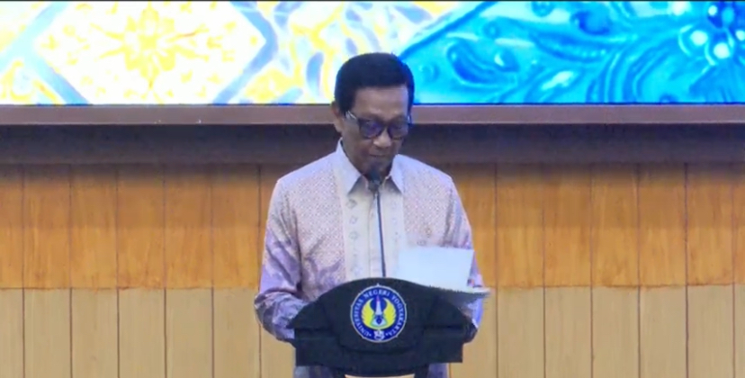 IMG_2359 - Mabur.co Man in a pink patterned shirt speaks at a podium with a microphone, reading from a folder, in a wood-paneled room with a blue screen backdrop.