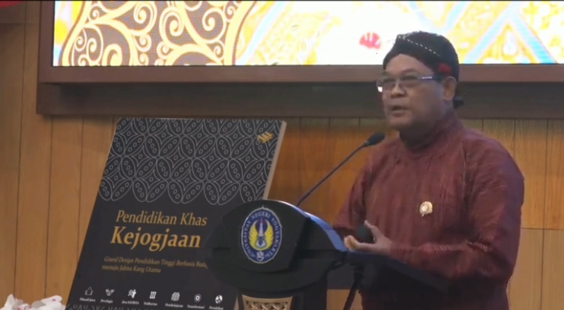 IMG_2362 - Mabur.co A man in a maroon traditional shirt speaks at a podium with a microphone, while a banner behind him reads'Pendidikan Khas Kejagoaan' and shows a patterned black design.