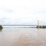 Suspension bridge with yellow towers spanning a wide, muddy river during high floodwaters. On- and off-ramps are barely visible on the banks.