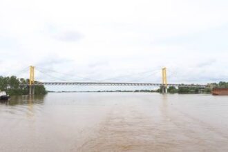 Suspension bridge with yellow towers spanning a wide, muddy river during high floodwaters. On- and off-ramps are barely visible on the banks.
