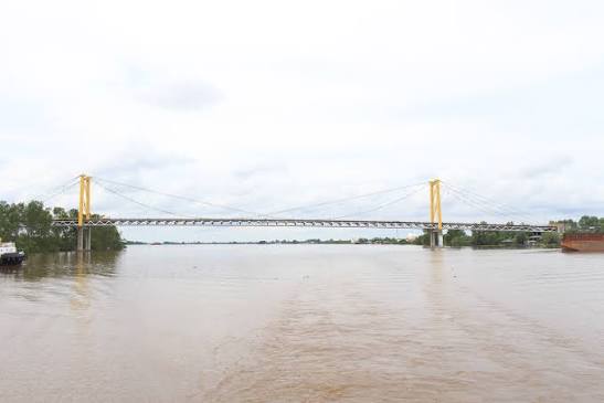 Suspension bridge with yellow towers spanning a wide, muddy river during high floodwaters. On- and off-ramps are barely visible on the banks.