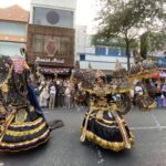 Dancers in elaborate black, gold, and purple costumes perform a street parade as a crowd watches along a city avenue with storefronts in the background.