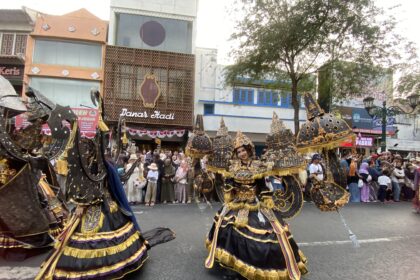 Dancers in elaborate black, gold, and purple costumes perform a street parade as a crowd watches along a city avenue with storefronts in the background.