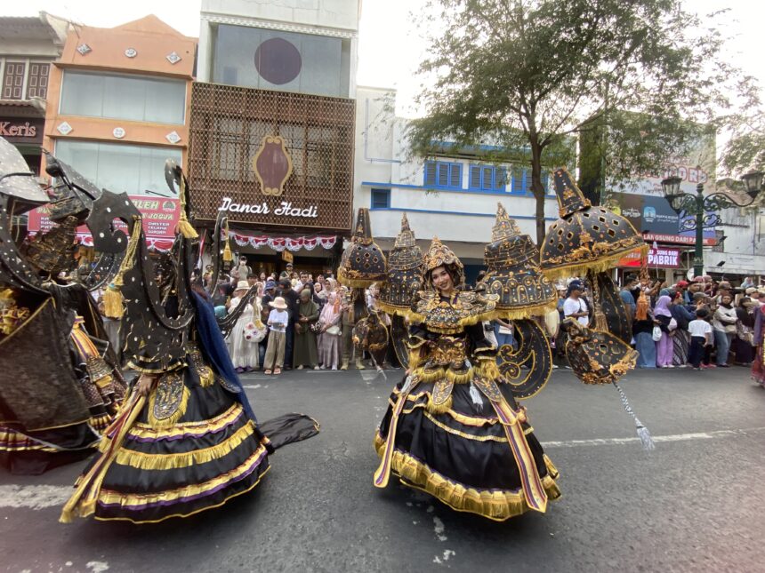 Dancers in elaborate black, gold, and purple costumes perform a street parade as a crowd watches along a city avenue with storefronts in the background.