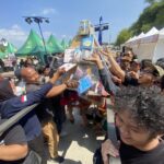 Crowded outdoor festival with people reaching toward a tall prize column wrapped in plastic under blue sky and tents behind them.