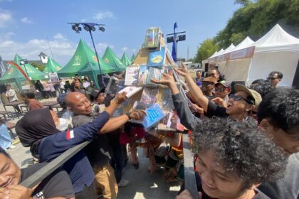Crowded outdoor festival with people reaching toward a tall prize column wrapped in plastic under blue sky and tents behind them.