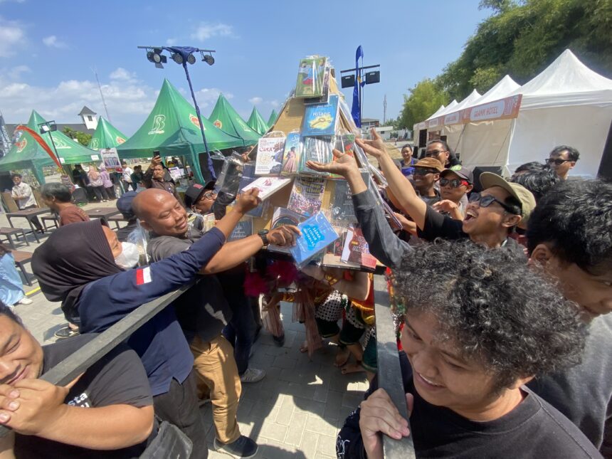 Crowded outdoor festival with people reaching toward a tall prize column wrapped in plastic under blue sky and tents behind them.
