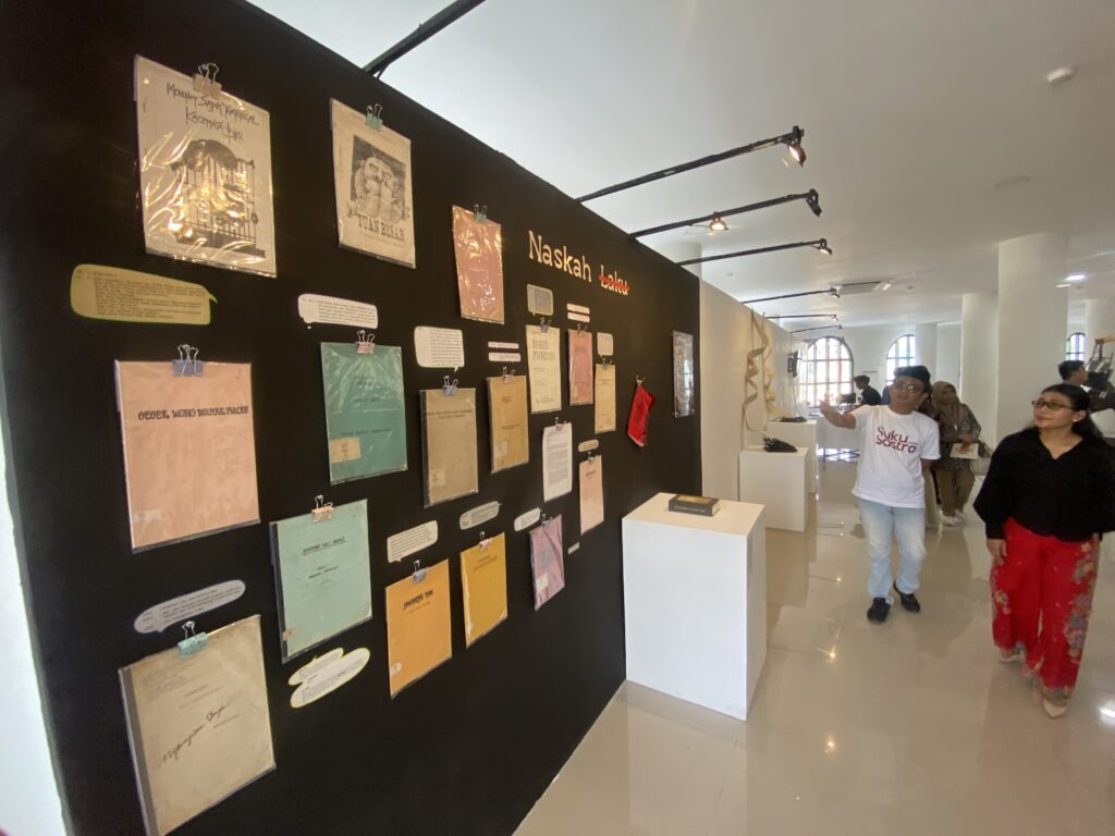 IMG_2502 - Mabur.co Black exhibition wall covered with colorful papers and notes clipped in place; visitors walk through a bright gallery space in the background.