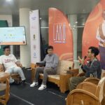 Panel discussion with three men seated on wooden chairs in a bright event space; one speaks into a microphone while another wears a white t-shirt with 'Suku Sastra' on it, backdrop features orange banners with artwork and logos.