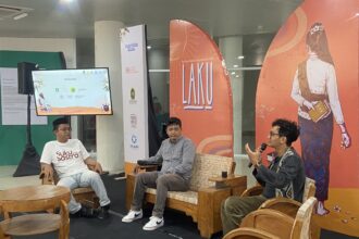 Panel discussion with three men seated on wooden chairs in a bright event space; one speaks into a microphone while another wears a white t-shirt with 'Suku Sastra' on it, backdrop features orange banners with artwork and logos.